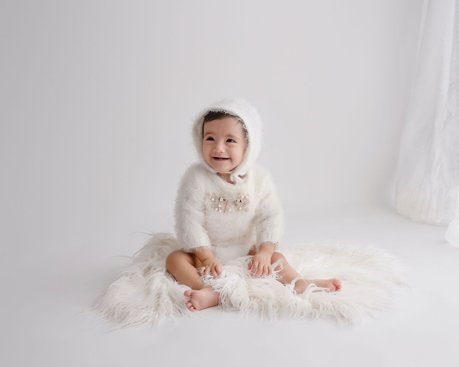 Baby wearing white outfit sits on soft fur backdrop in bright minimalist studio setting.