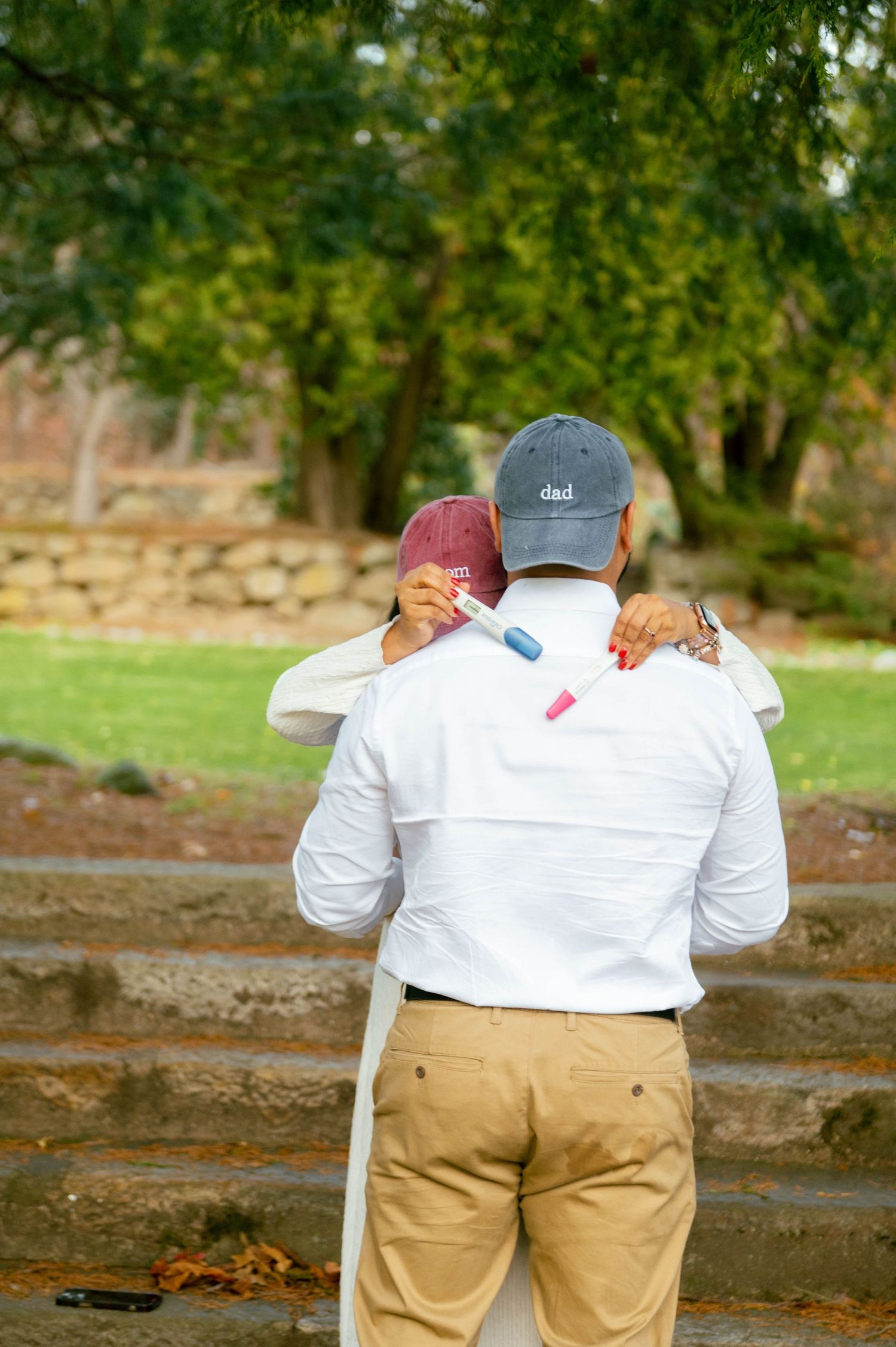 Couple embracing outdoors, holding pink and blue pregnancy test markers, man wearing 'dad' cap, gender reveal moment.