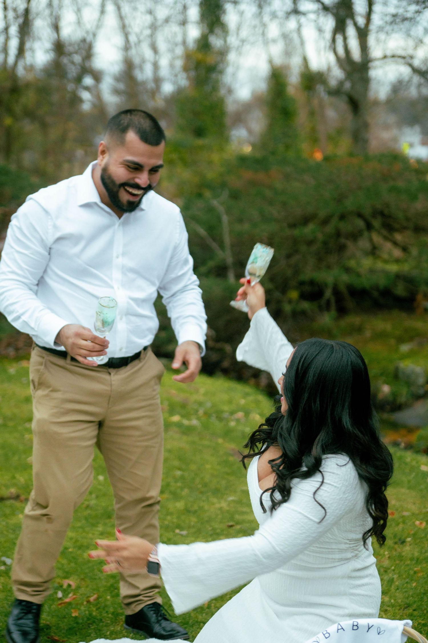 Couple in white outfits toasting with champagne glasses outdoors in a garden setting.