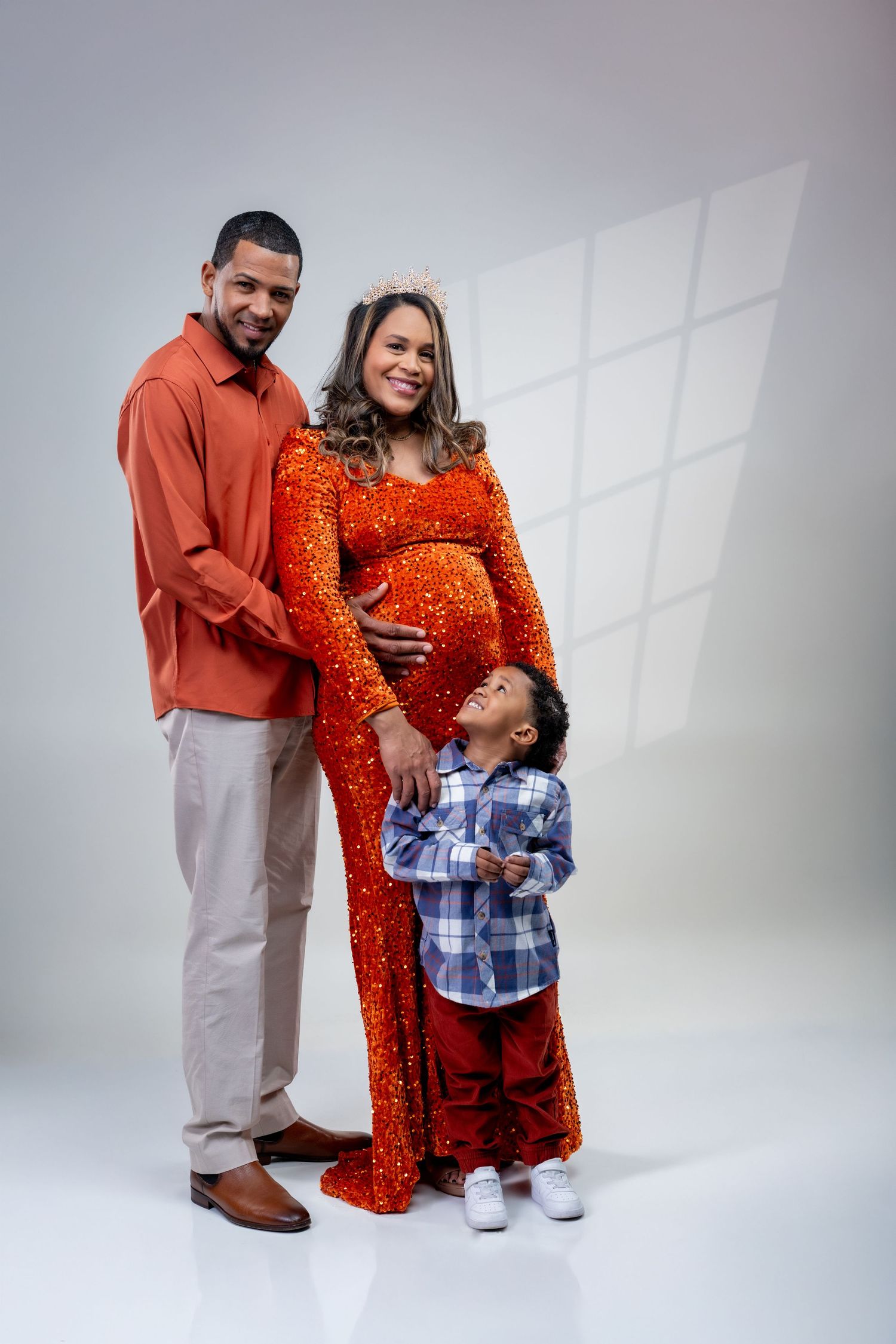 A pregnant woman in a red sequin gown and crown poses with her husband and young son in a studio photo.