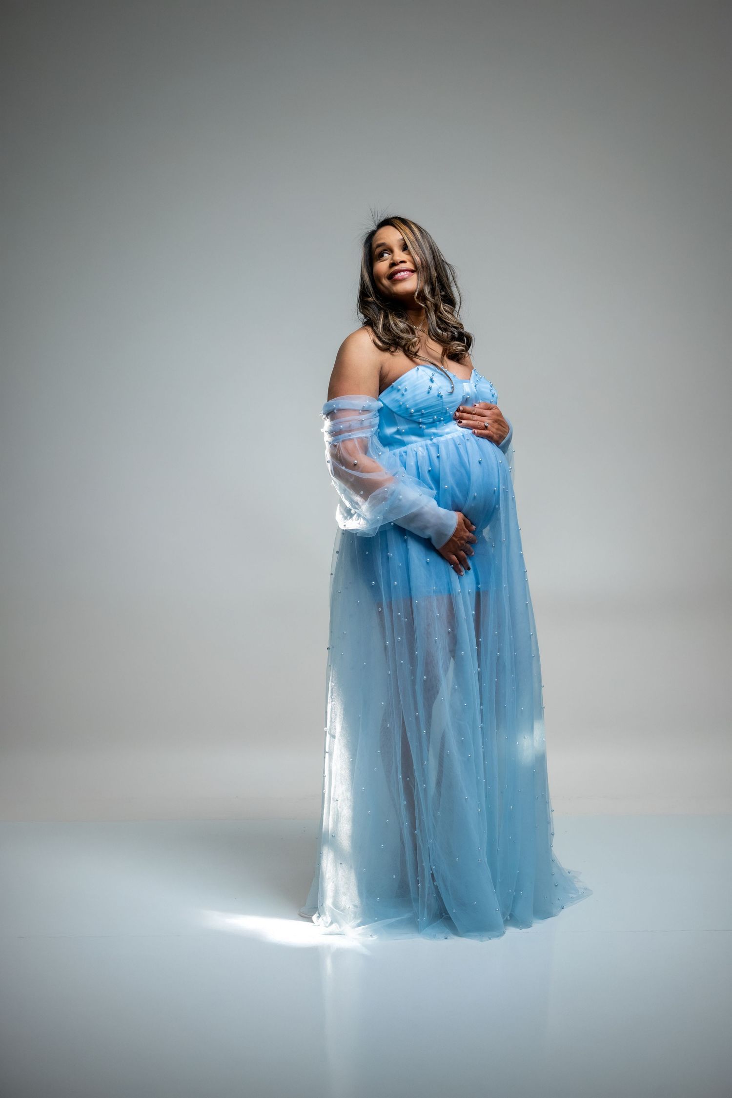 Pregnant woman in flowing blue tulle gown cradling baby bump, smiling softly in professional maternity photo shoot.