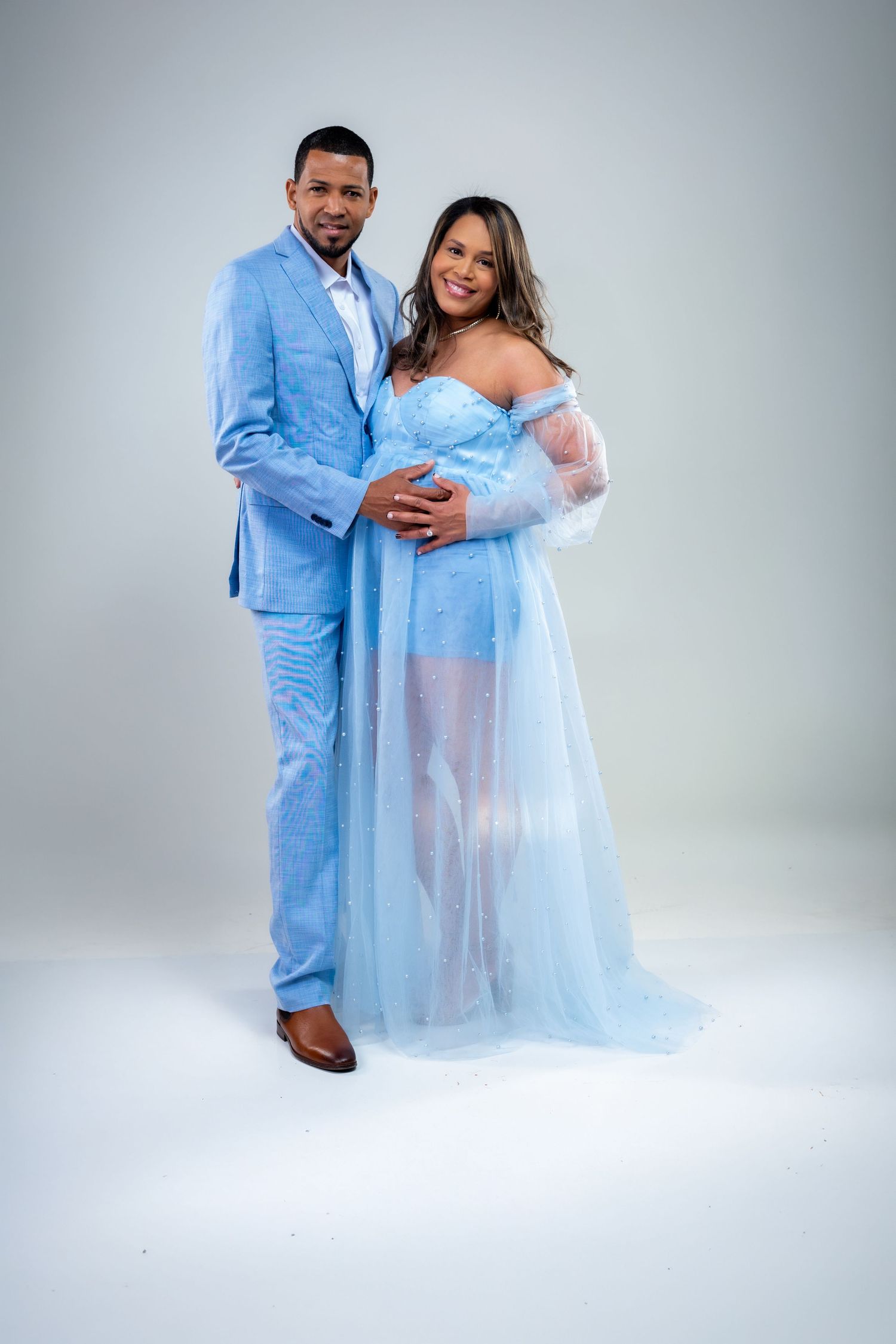 Couple in matching light blue outfits posing together, pregnant woman in tulle gown, man in blue suit, studio photo.