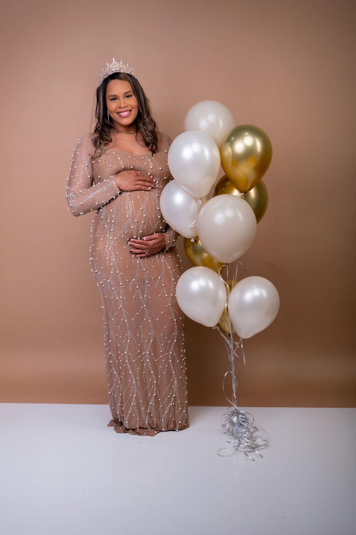 Smiling pregnant woman in beaded gown and crown holding belly beside gold and white balloons.