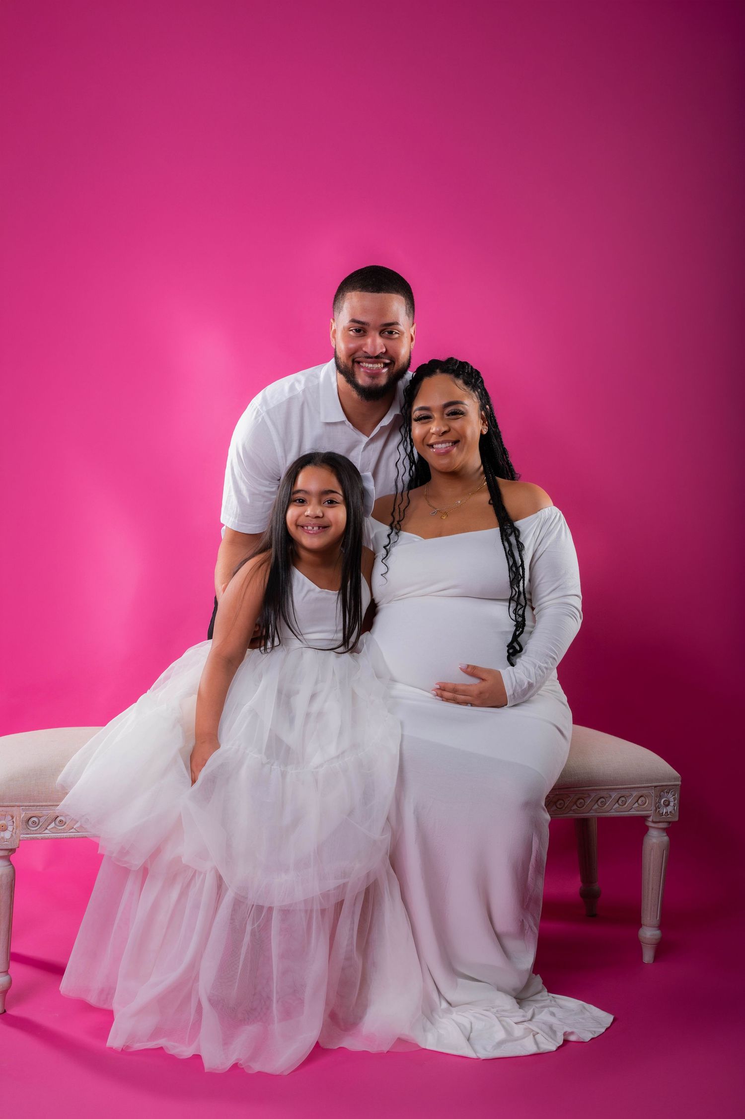 A family maternity photo with a pregnant woman, man, and young girl in white outfits against a bright pink background.