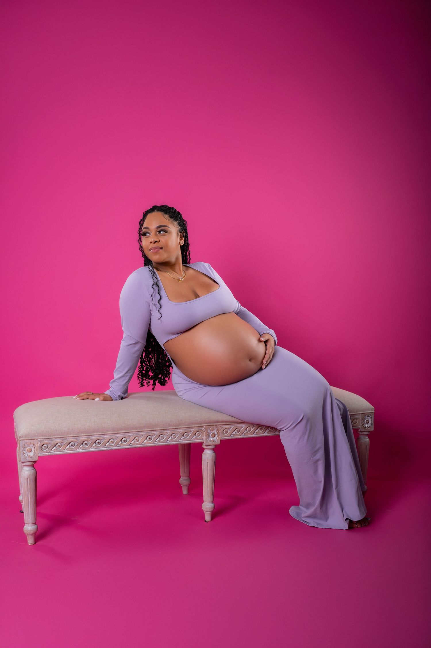 Pregnant woman in lavender dress poses on elegant bench against vibrant pink background, cradling her baby bump.