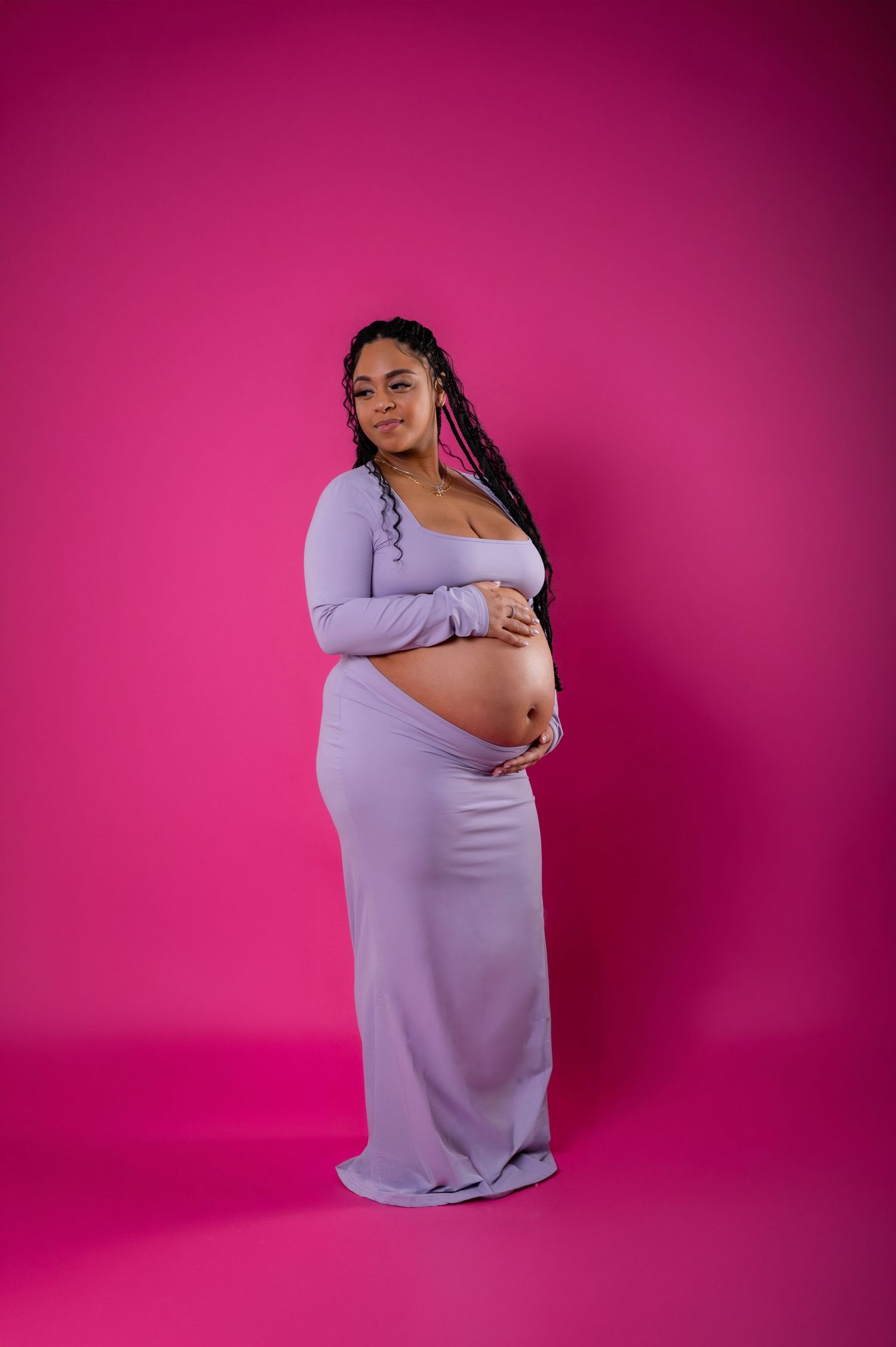Pregnant woman in lavender maxi dress holding baby bump against vibrant pink background.