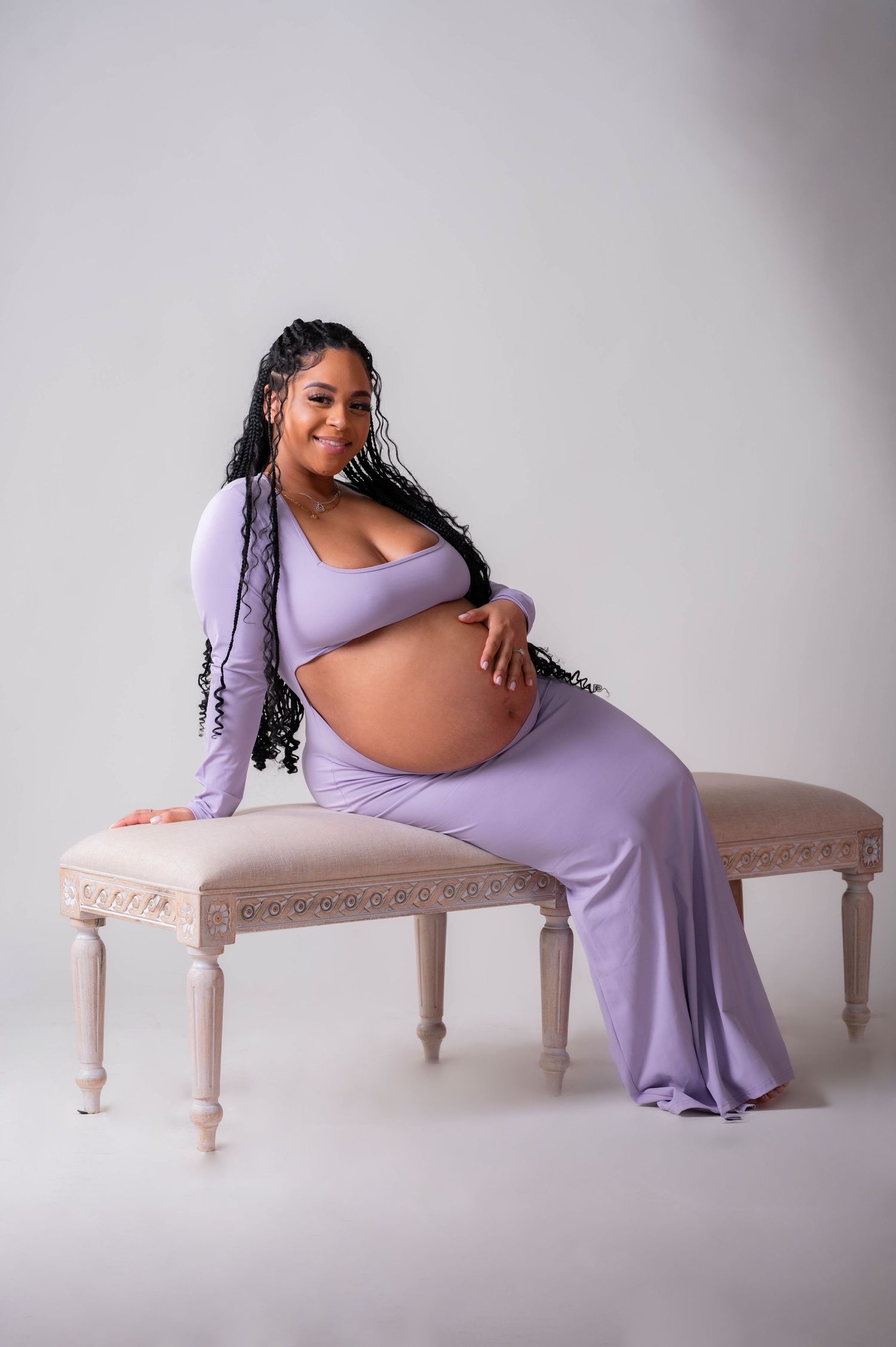 Pregnant woman in lavender maternity outfit sitting on elegant bench, cradling baby bump, with long braids.