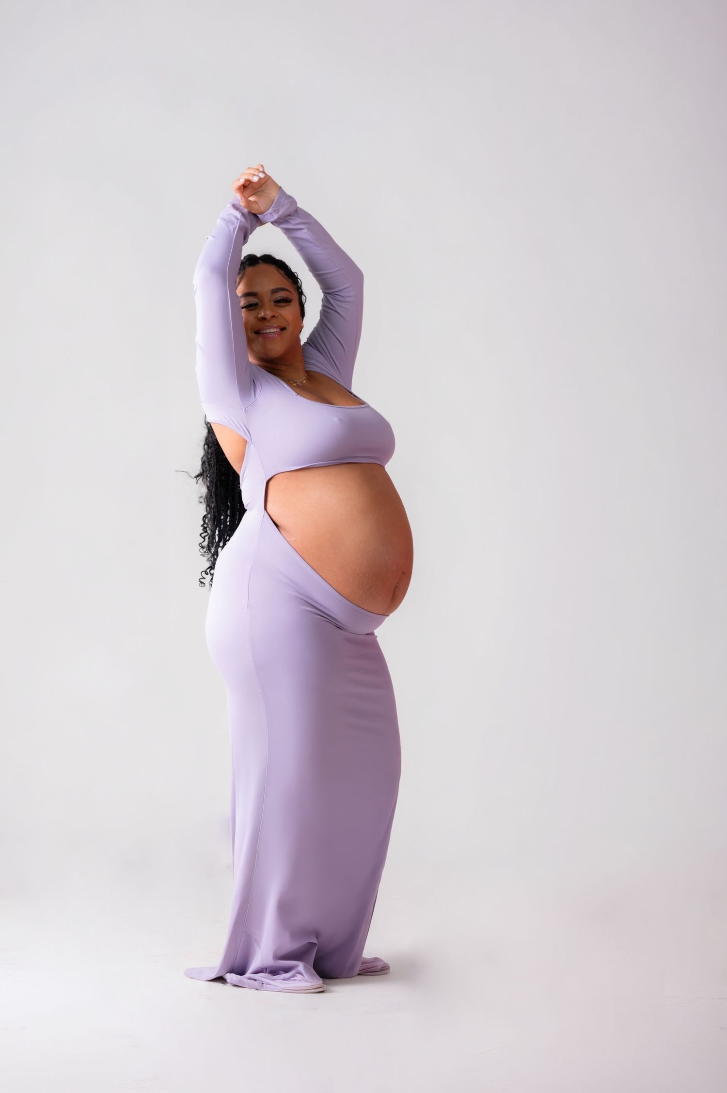 Pregnant woman in lavender cutout maternity gown posing with arms raised on white background.