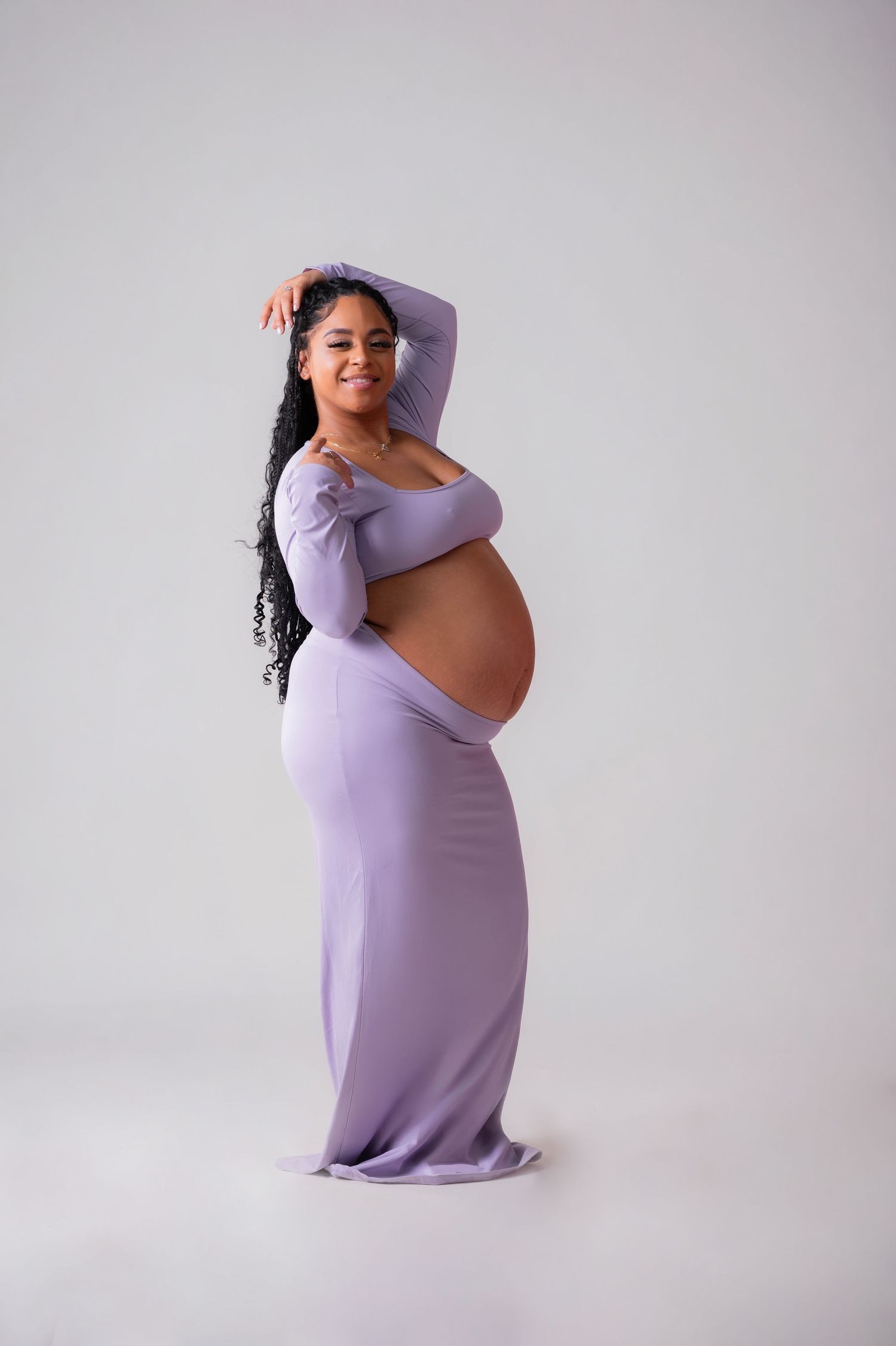 Smiling pregnant woman in purple off-shoulder maternity dress cradling baby bump in studio.