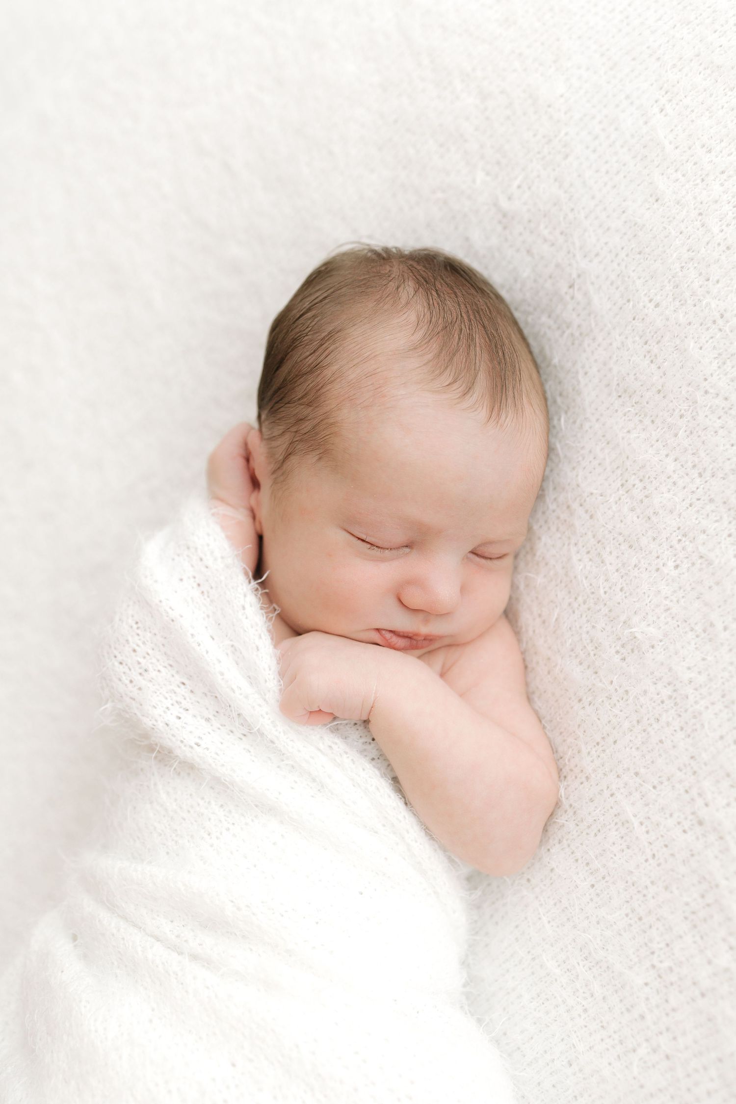 Sleeping newborn wrapped in soft white blanket on textured background.