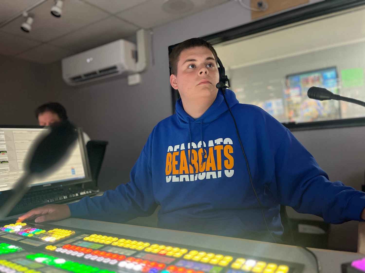 Student in blue hoodie operates broadcasting equipment in studio setting.