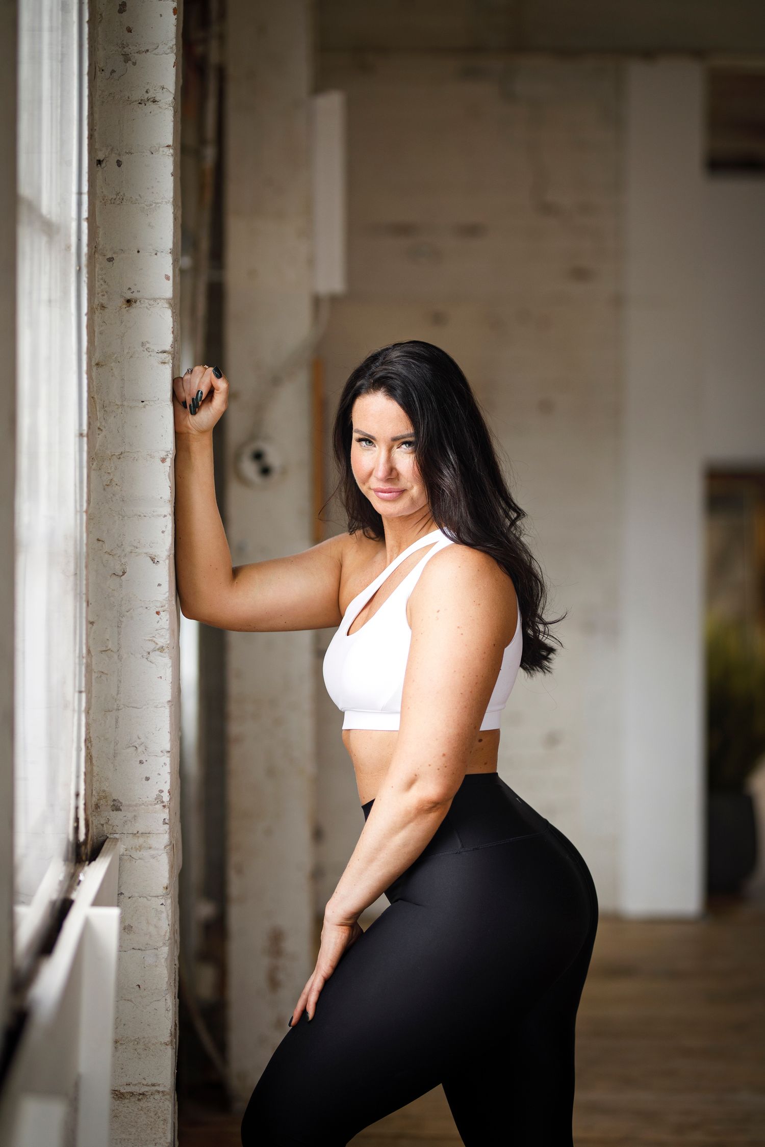 Athletic woman in workout attire poses confidently against white brick wall during fitness photoshoot.