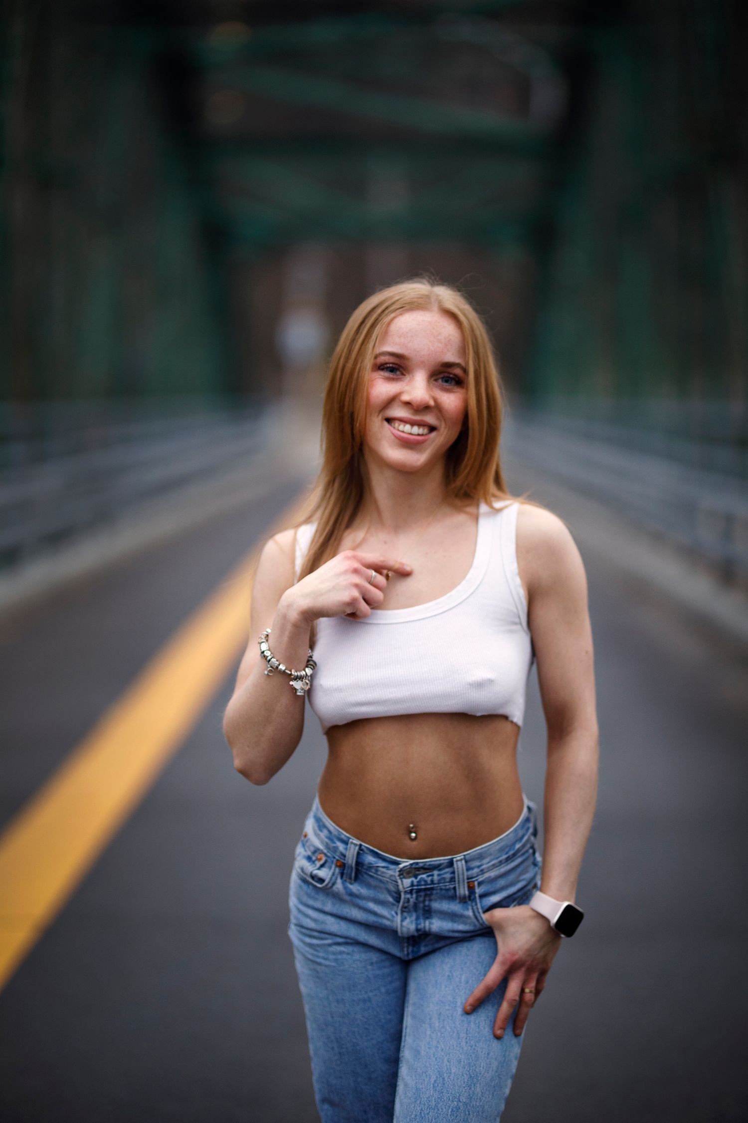 A woman in a white crop top and jeans poses on an empty road with trees and railings in the background.