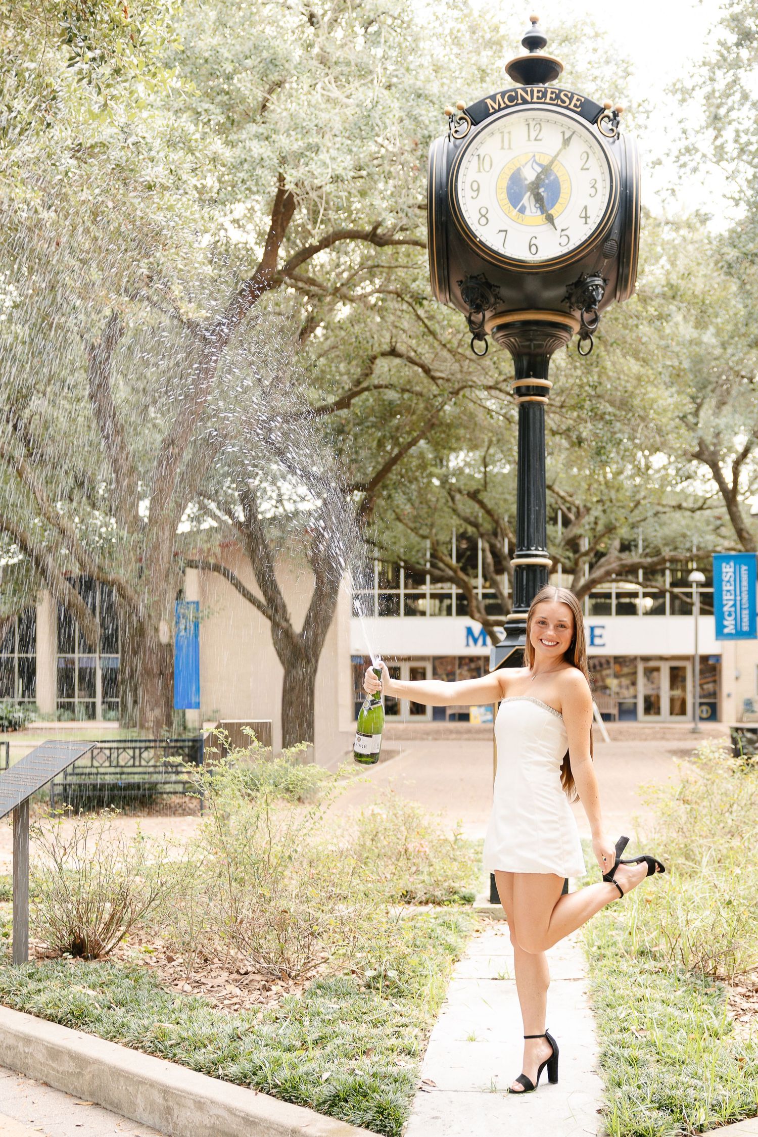 Young woman in white dress celebrating near McNeese State University clock, popping champagne bottle outdoors.
