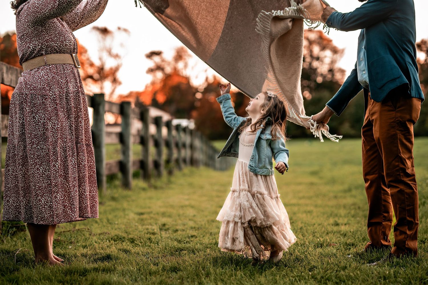 Child wearing layered dress swings between parents' hands in a golden sunset field with wooden fence in background.