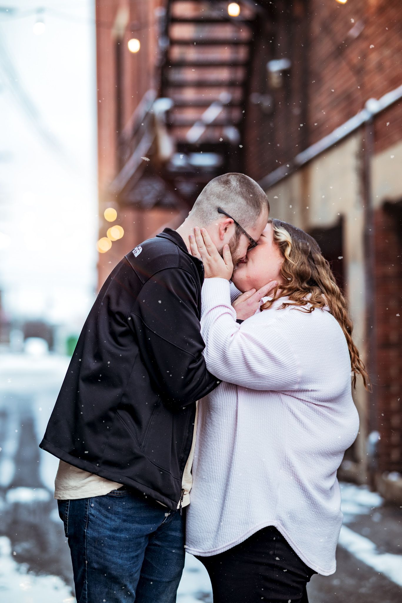 A couple shares a romantic kiss on a snowy winter day in front of a brick building with fire escapes.