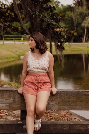 Relaxed pose on wooden bench by lake wearing summer outfit with coral shorts.