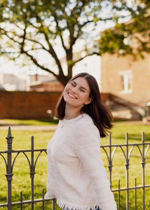 Person in white sweater smiling against iron fence and autumn backdrop.