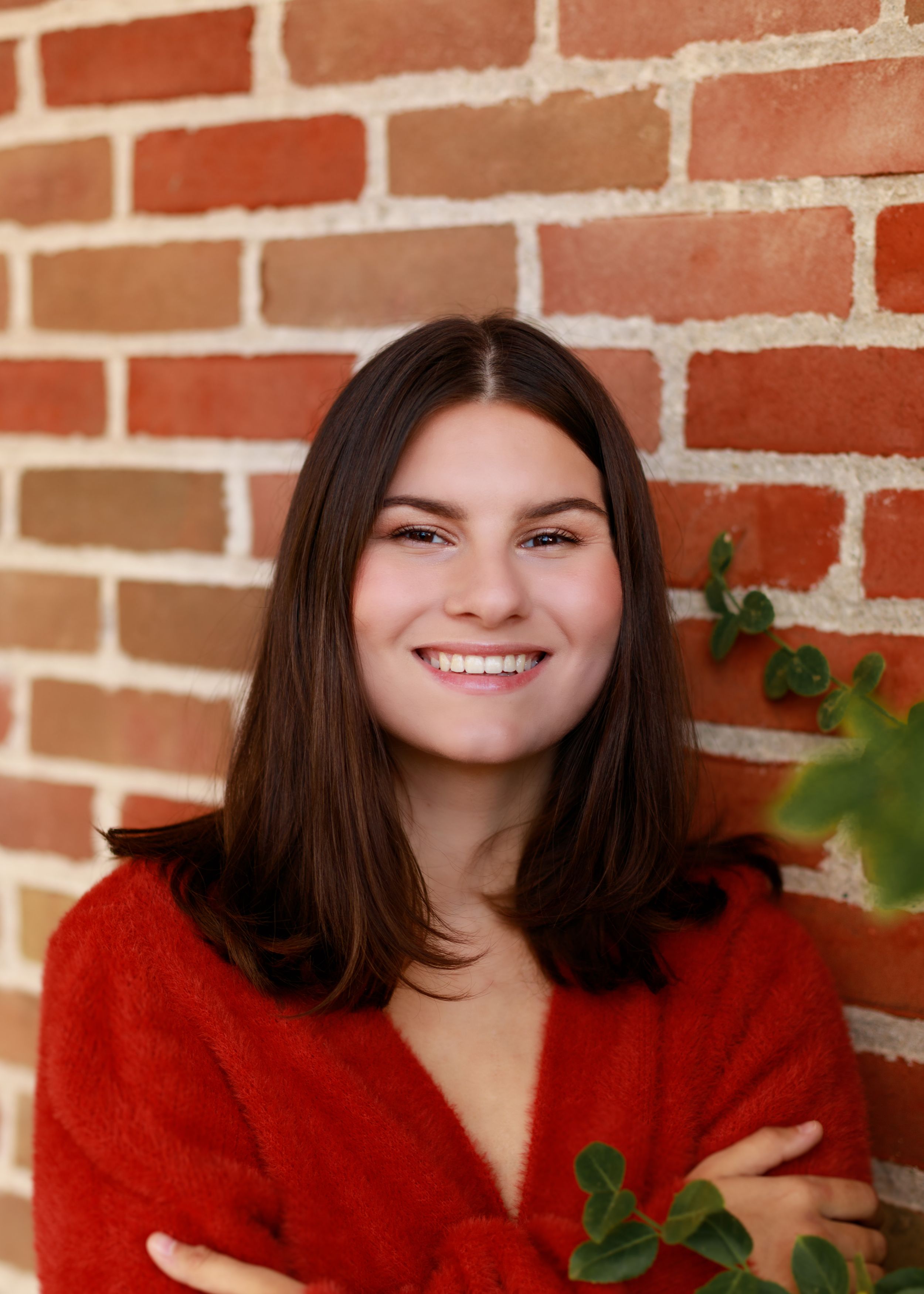 Portrait series against a red brick wall featuring a bright red sweater and natural greenery.