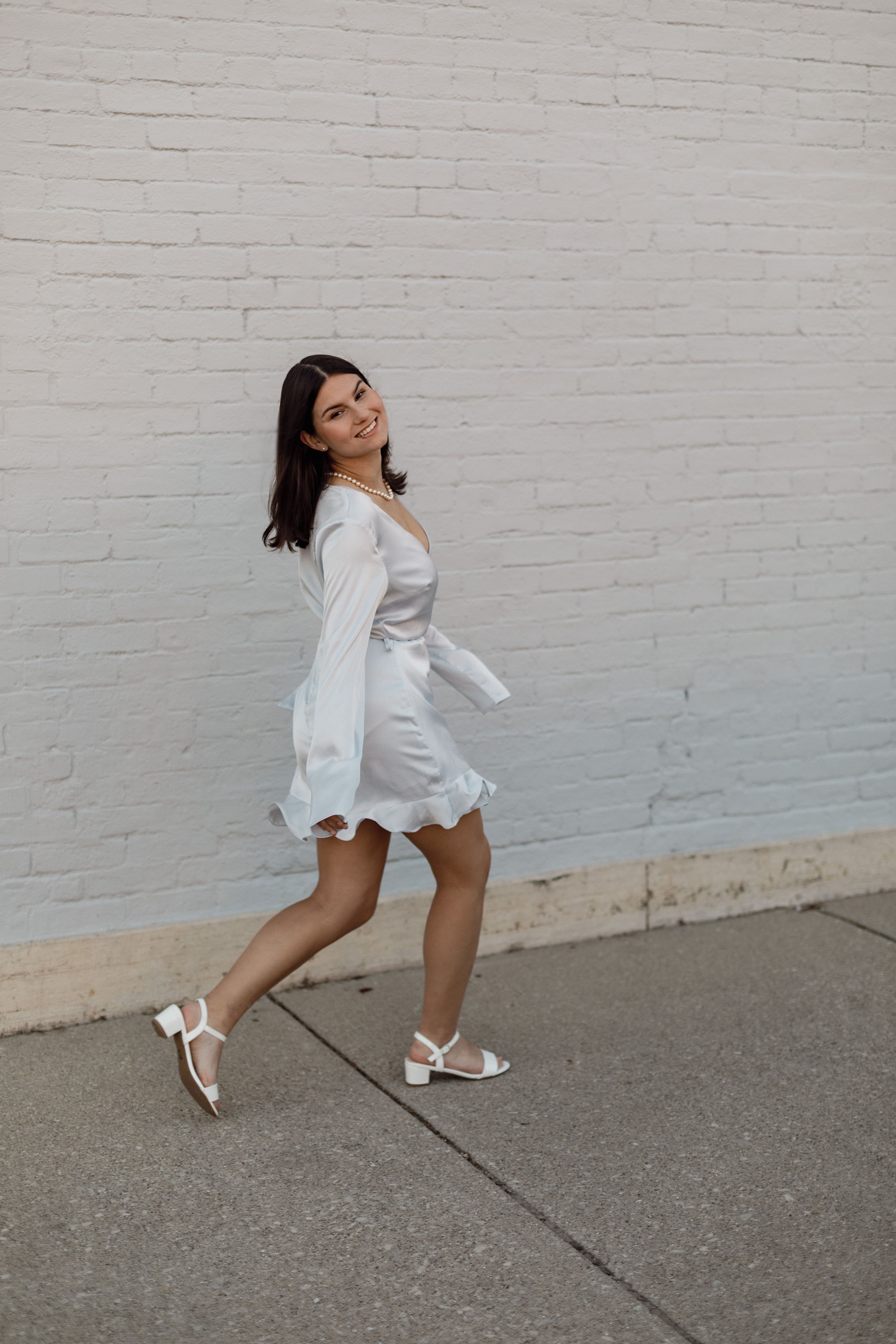 Playful dance-like poses in a white dress against a painted brick wall.