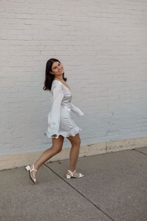 Playful dance-like poses in a white dress against a painted brick wall.