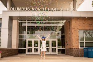 Graduate throwing confetti in celebration outside high school entrance.
