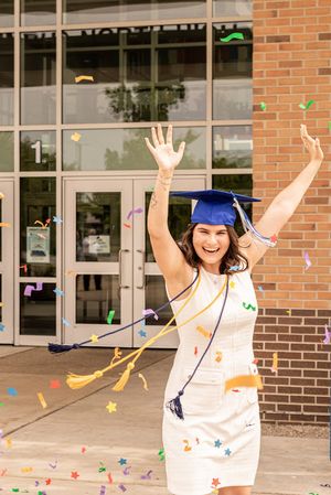 Graduate in white dress and blue cap celebrating with confetti outside brick building.