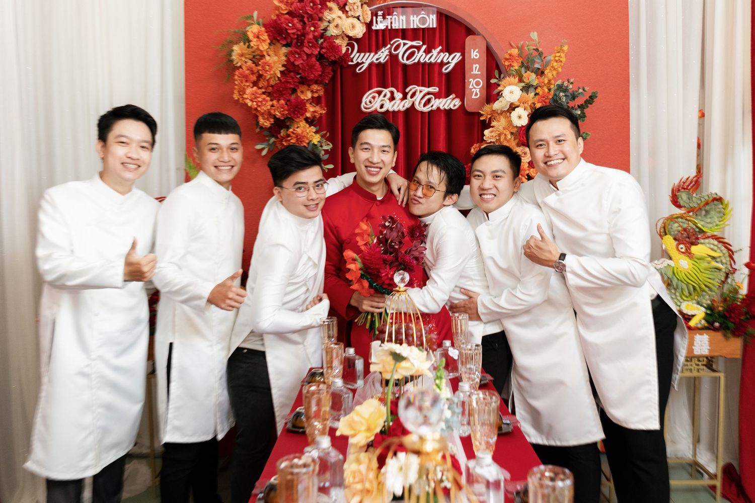 Festive red and gold wedding banquet table with group in white attire.