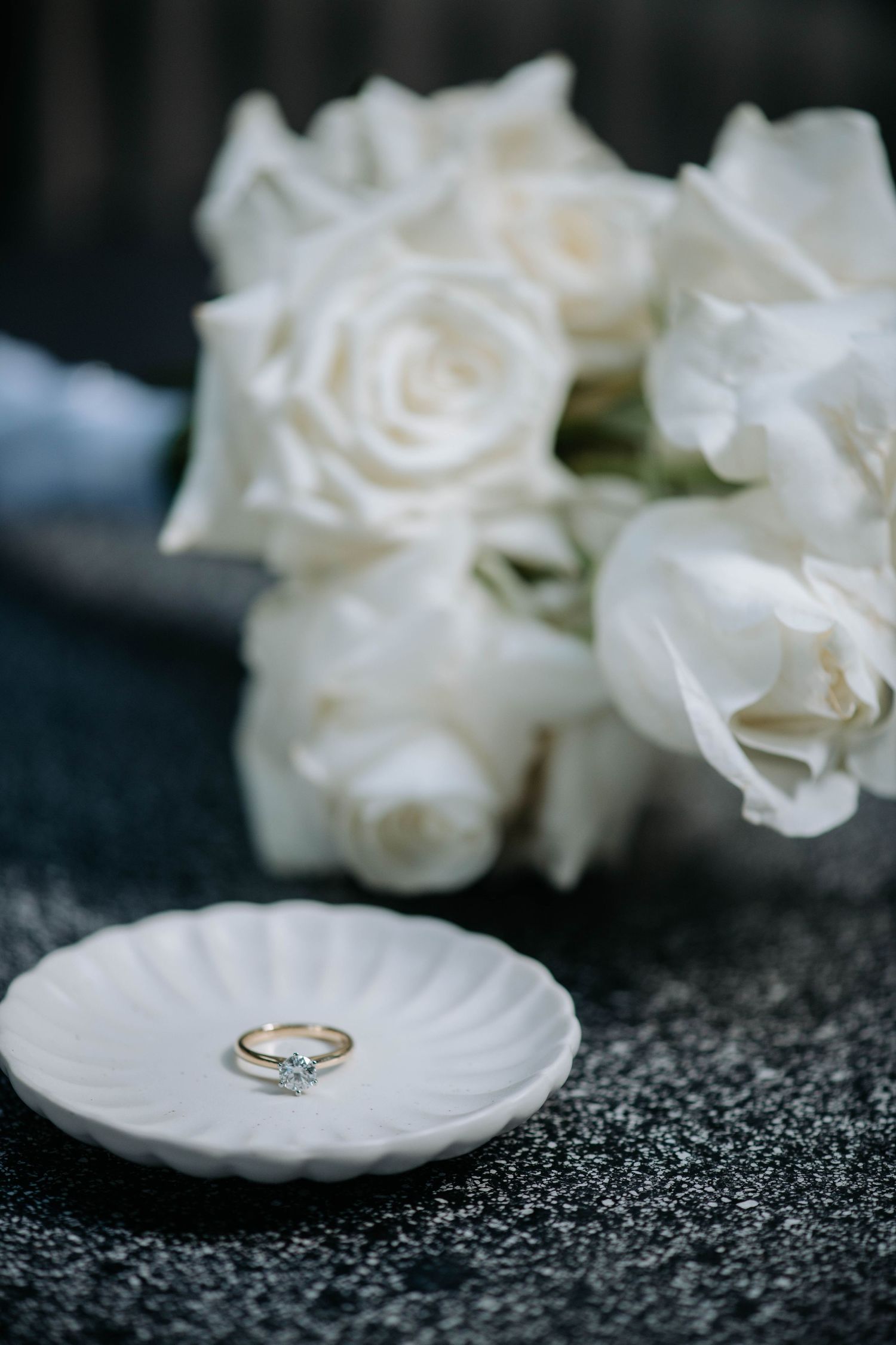 White roses and wedding rings arranged on a scalloped dish against dark background.