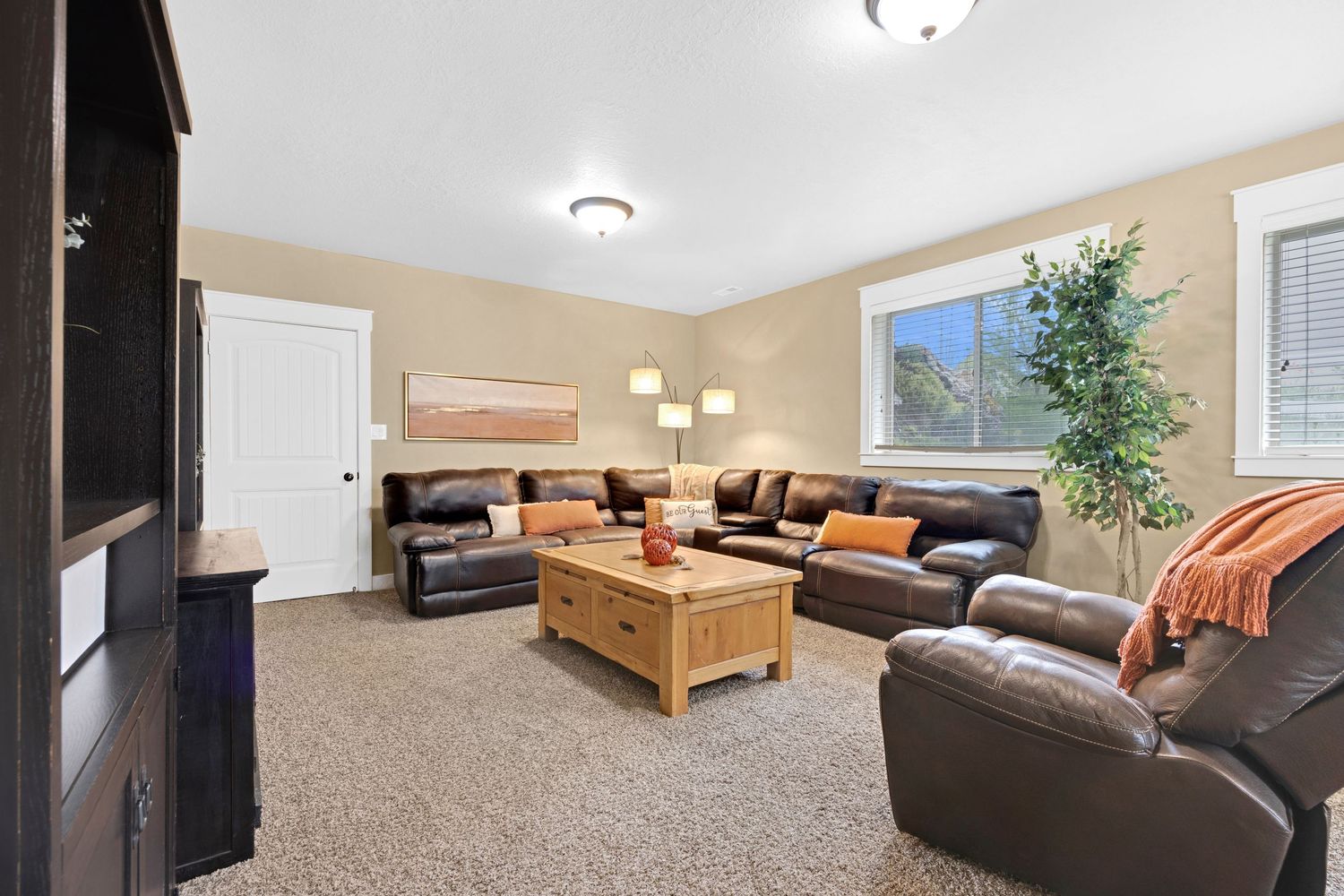 Cozy carpeted living room with leather sectional sofas, wooden coffee table, and large window with natural light.