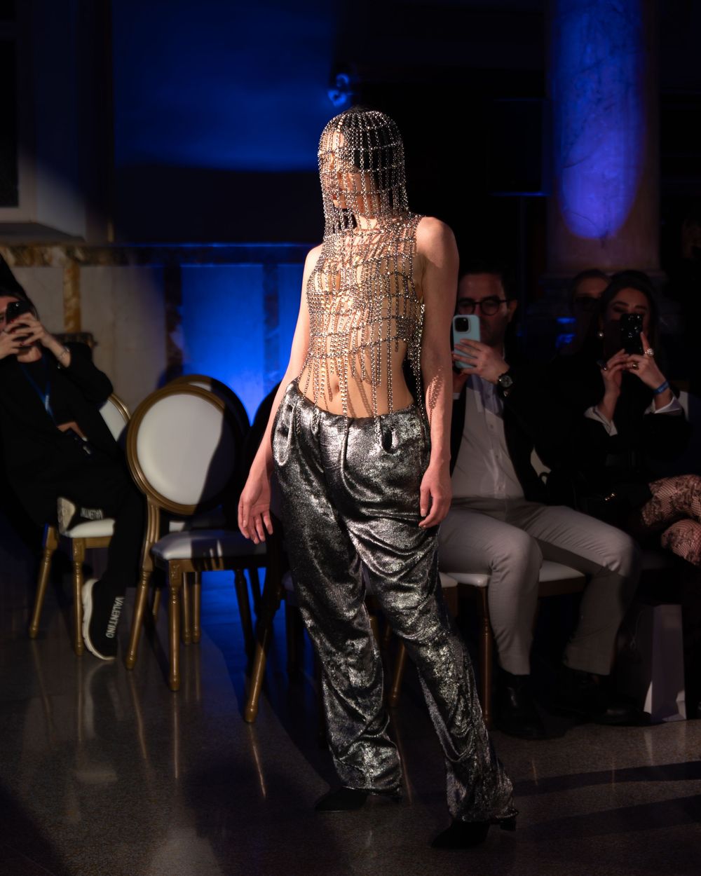 Fashion model walks runway in metallic pants and avant garde headpiece at blue-lit fashion show with audience.
