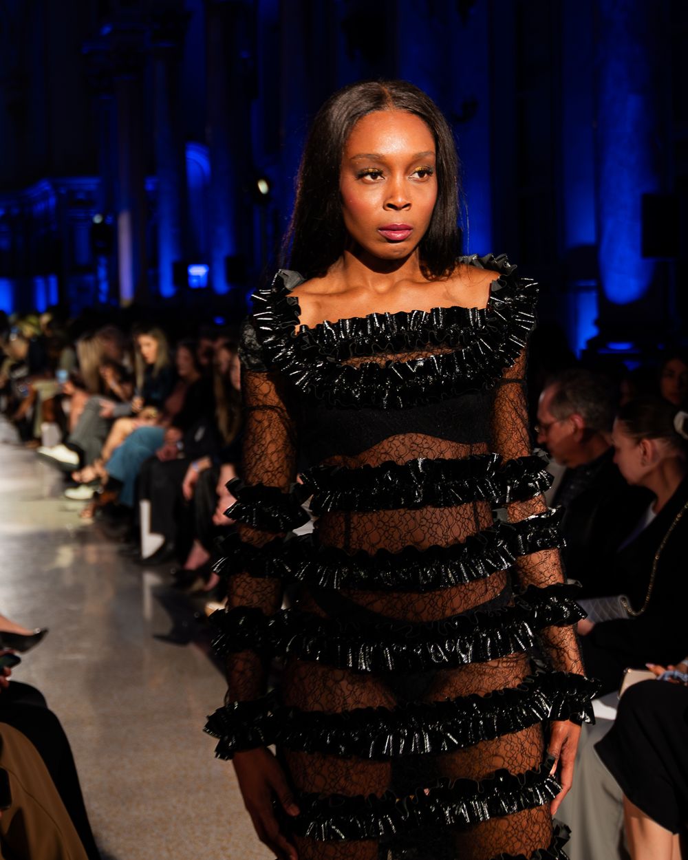 Model walks runway in black sheer dress with dramatic ruffled tiers during evening fashion show.