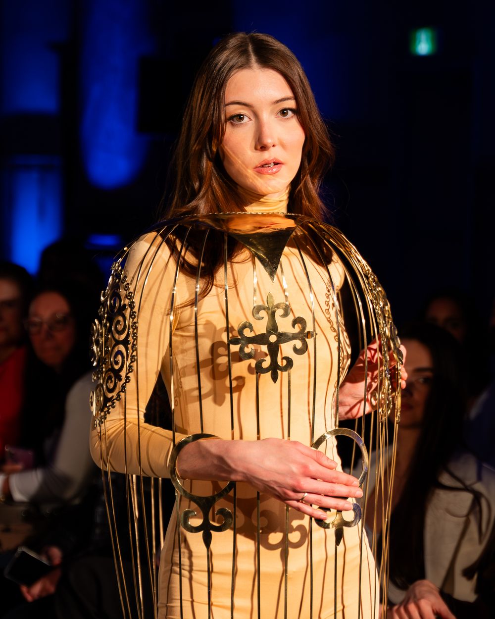 A model walks the runway wearing a cream-colored dress with ornate embellishments during a fashion show presentation.