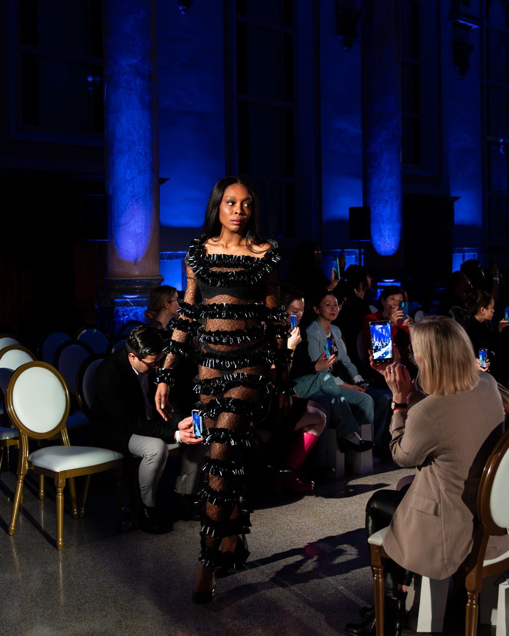 A fashion show model walks the runway in a black tiered lace dress with dramatic sheer panels under blue lighting.