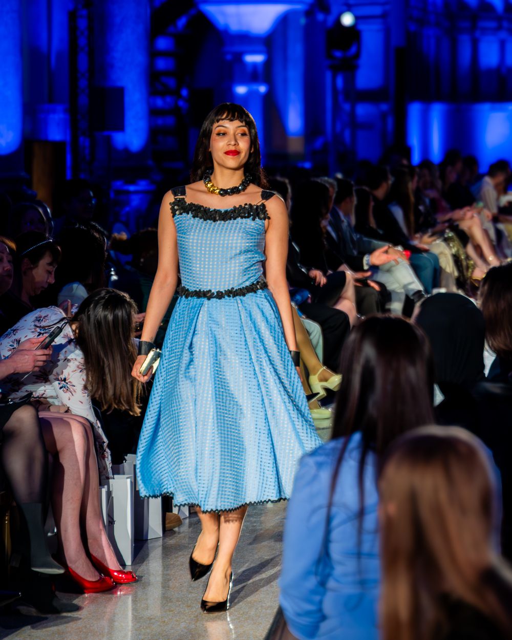 Model showcases vintage-inspired light blue gingham dress with black trim at fashion show runway.