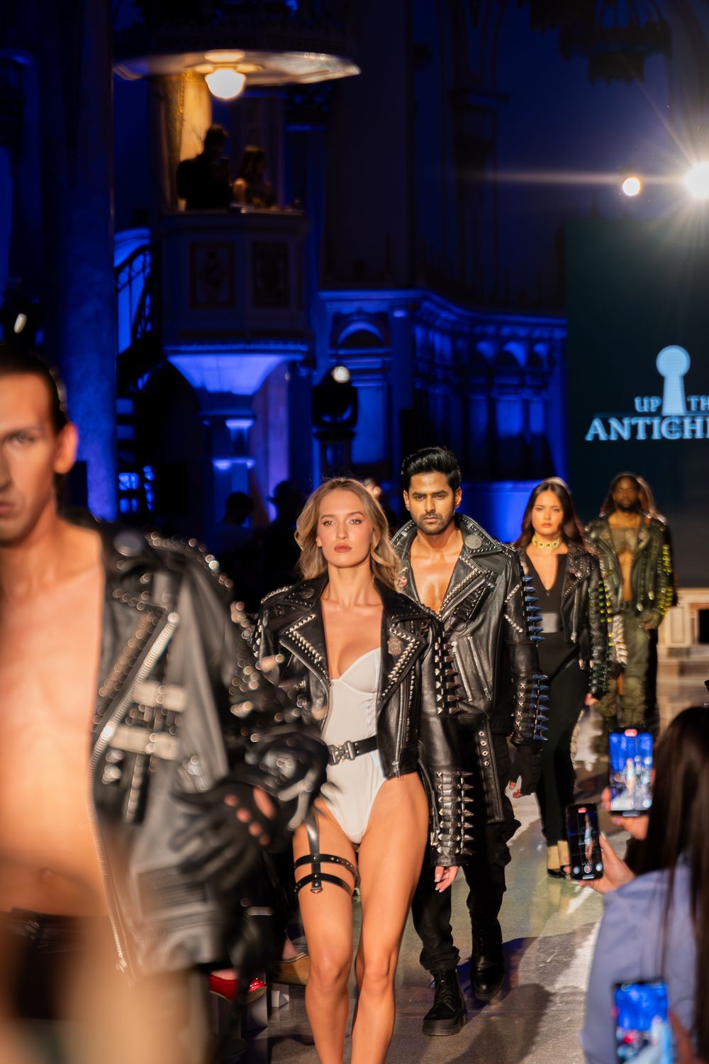 Models walk the catwalk in edgy leather and metallic outfits during a nighttime fashion show with blue lighting.