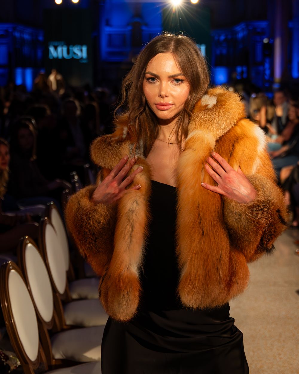 Fashion model walks runway in luxurious red-orange fur coat over black dress at glamorous evening fashion show.