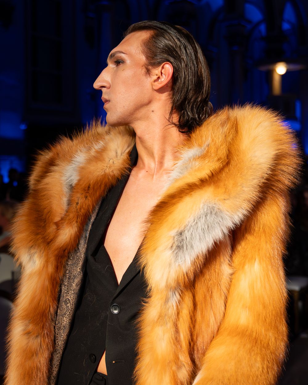 Model wearing luxurious orange fur coat walks fashion runway under dramatic blue lighting.