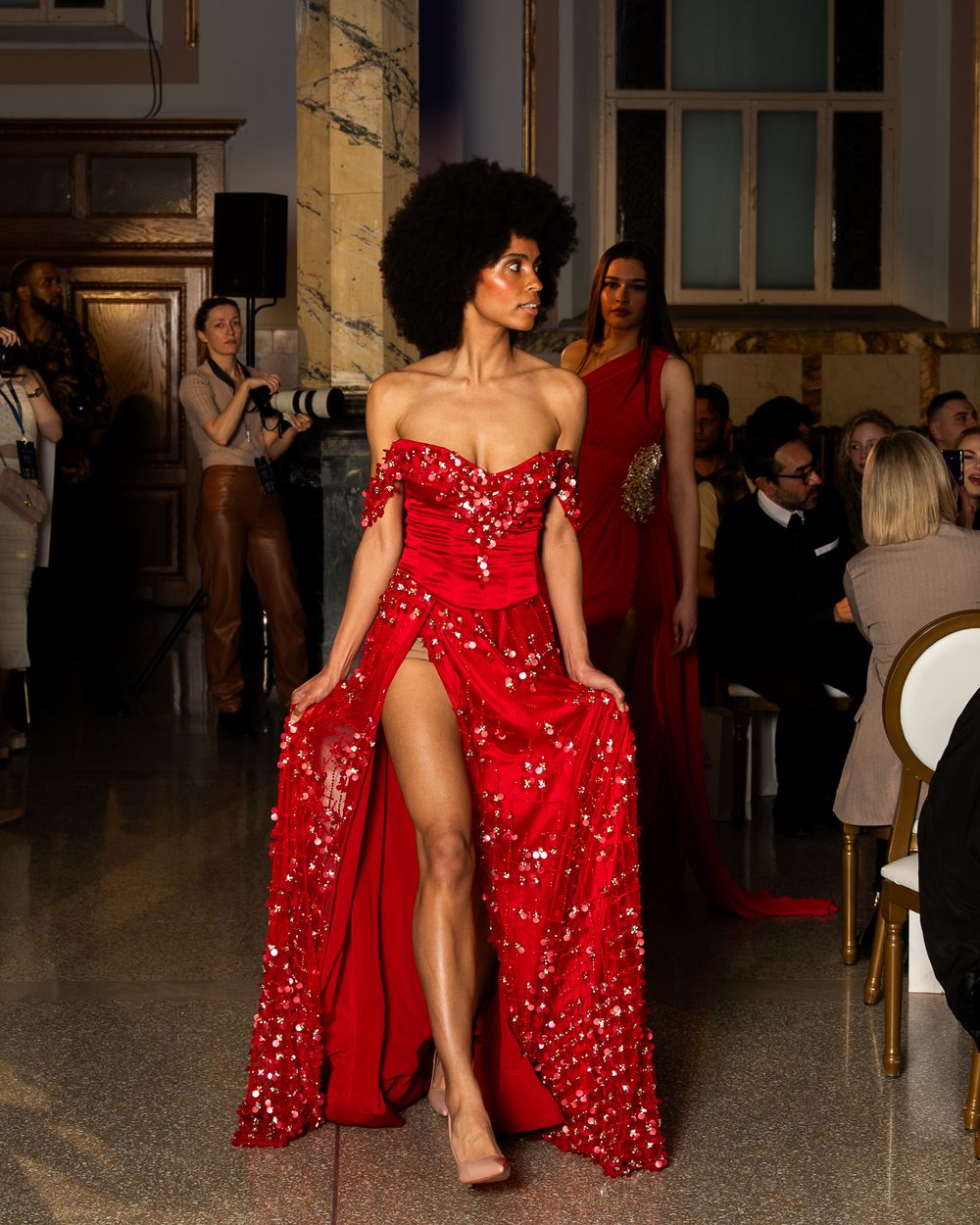 Fashion model wearing a stunning red sequined gown with high slit and off-shoulder design walks down an elegant hallway.