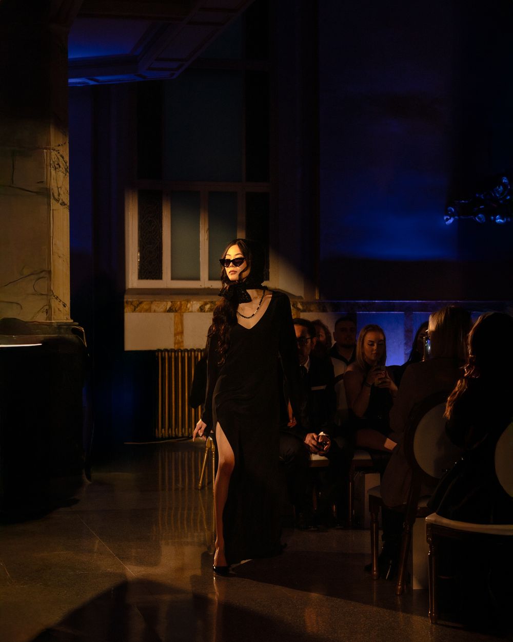 Model walks dramatic dark runway in black evening gown during moody blue-lit fashion show presentation.