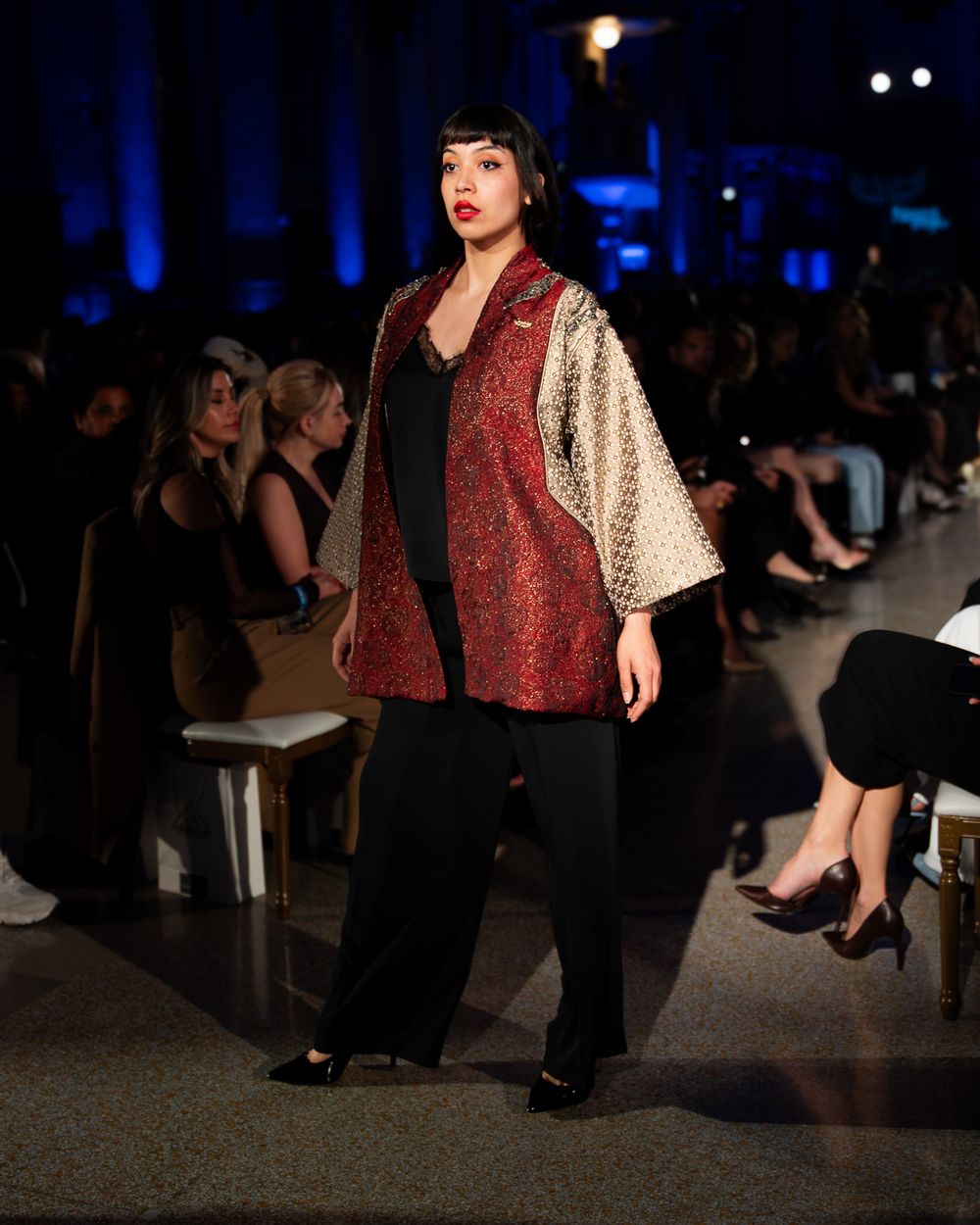Plus size model walks runway in red and cream contrast kimono jacket over black outfit at fashion show event.