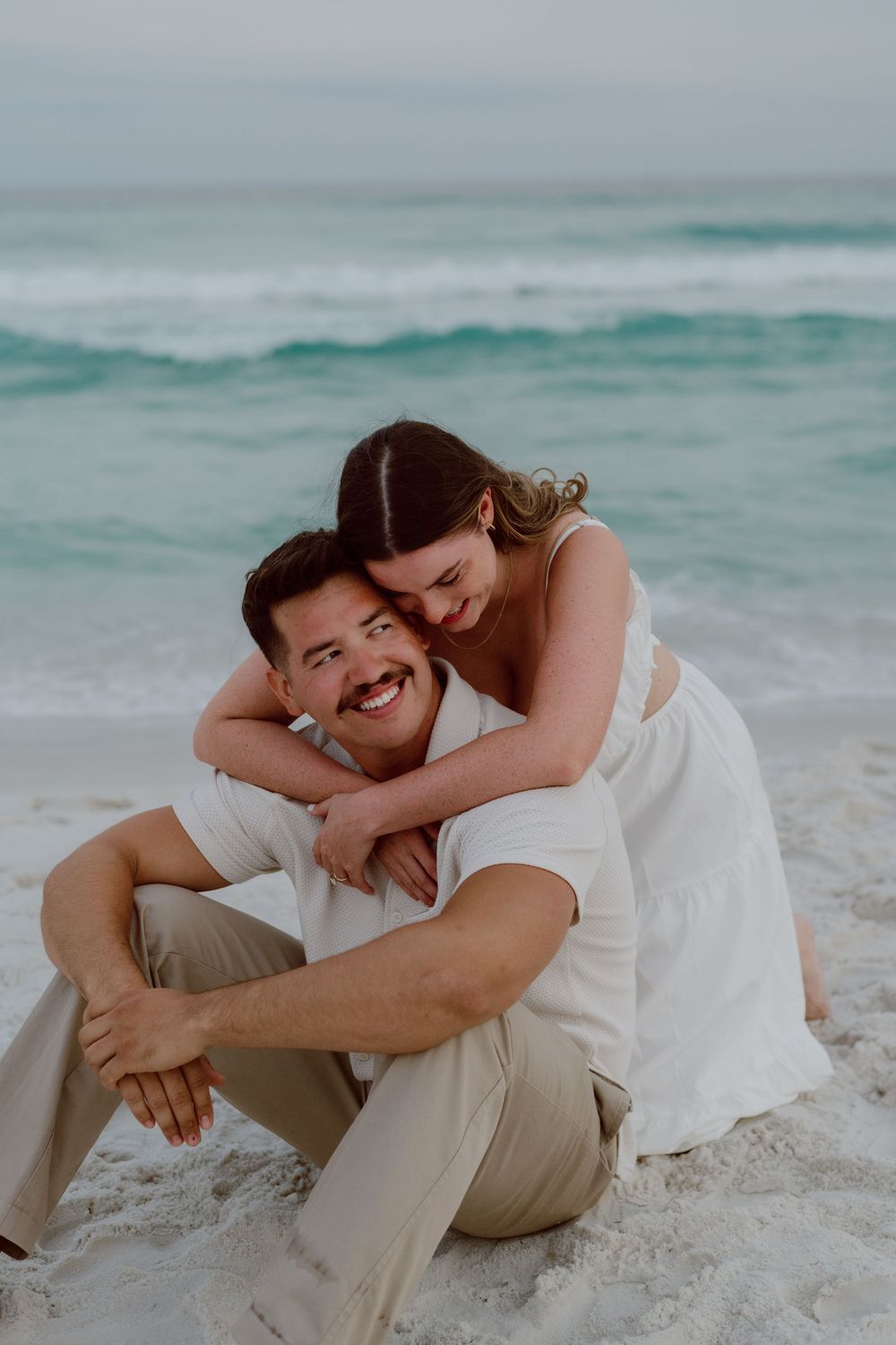 Couple embracing and laughing together on a sandy beach with ocean waves in the background.