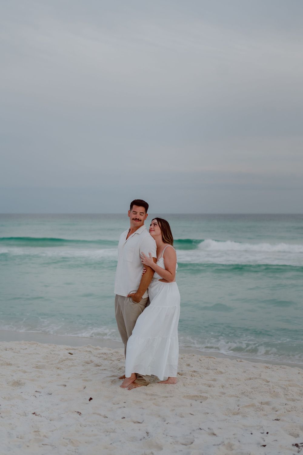 A couple embraces on a white sandy beach with turquoise waves rolling in under an overcast sky.
