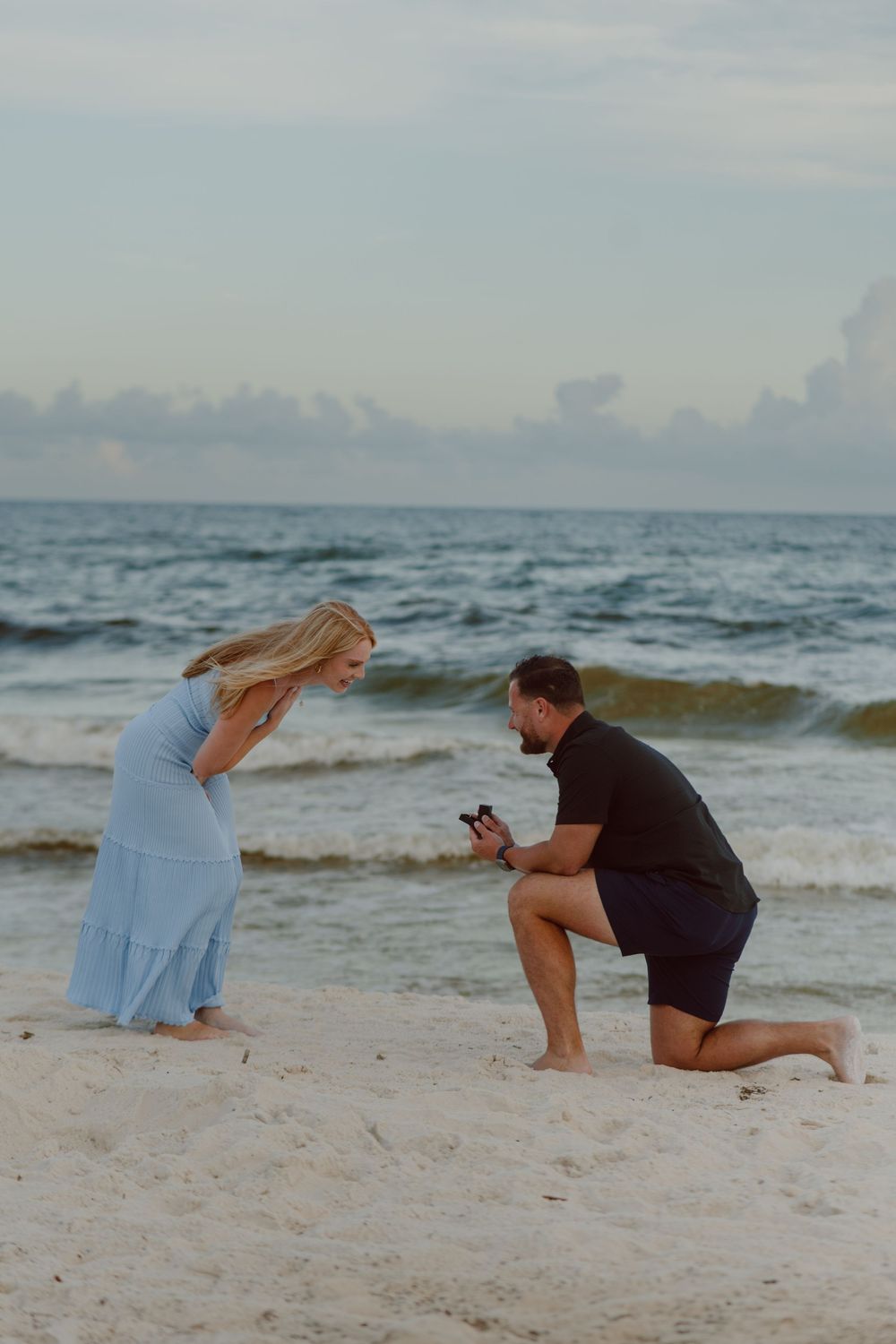 A couple performs a surprise beach proposal during sunset on a sandy shoreline.
