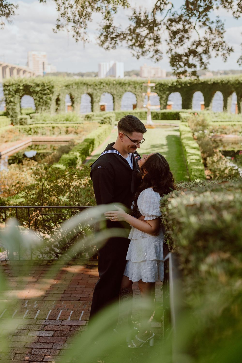 A romantic couple shares an intimate moment in a garden setting with brick walls and trees in the background.