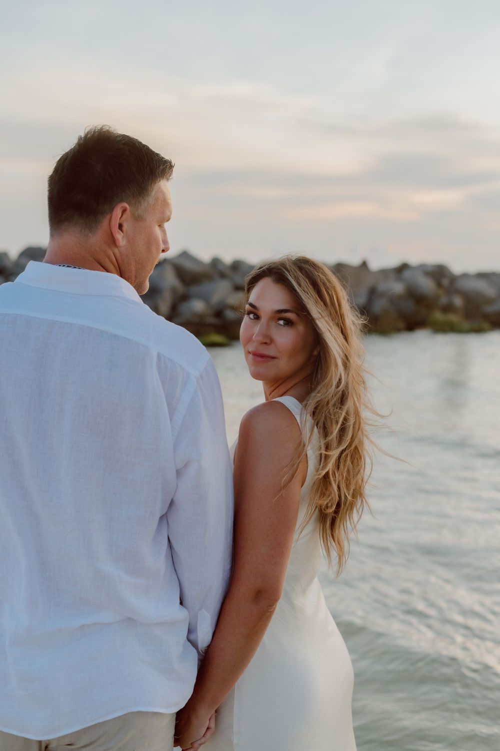Couple in white attire share romantic moment at sunset along rocky coastline near calm ocean waters.