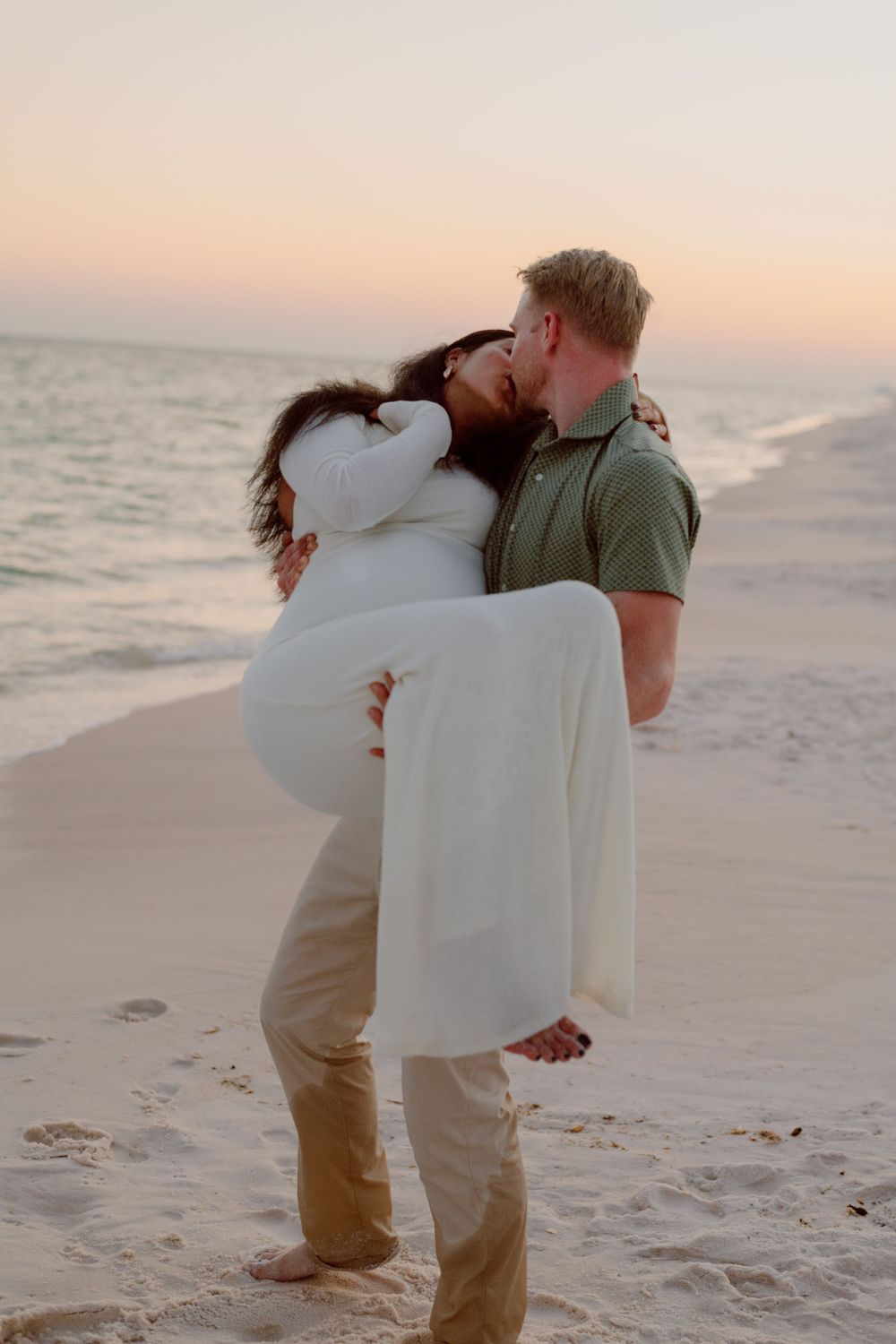 Romantic beach scene with person carrying partner in white dress at sunset.