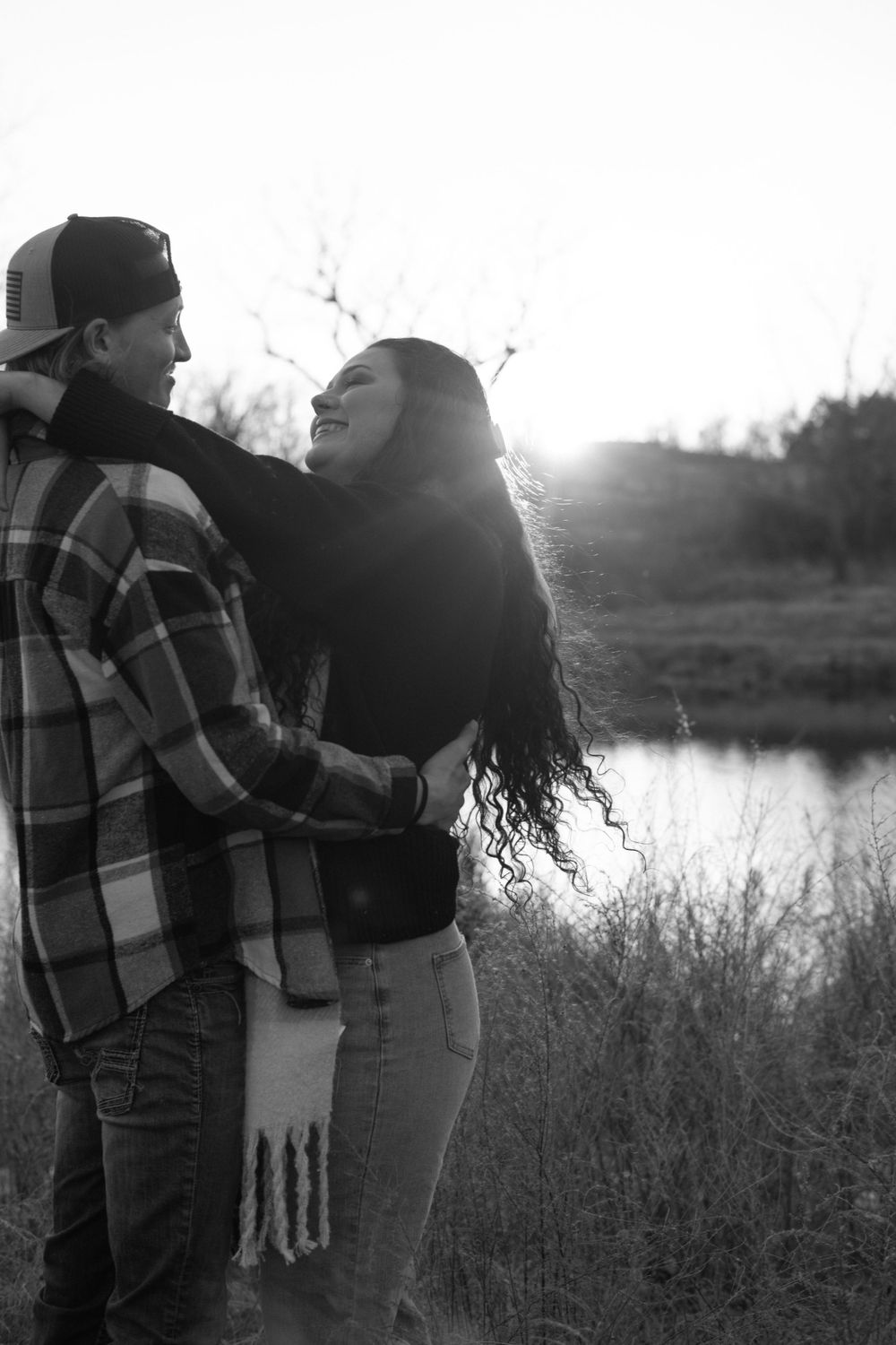 Romantic couple embracing by a lake at sunset wearing casual attire in black and white photograph.