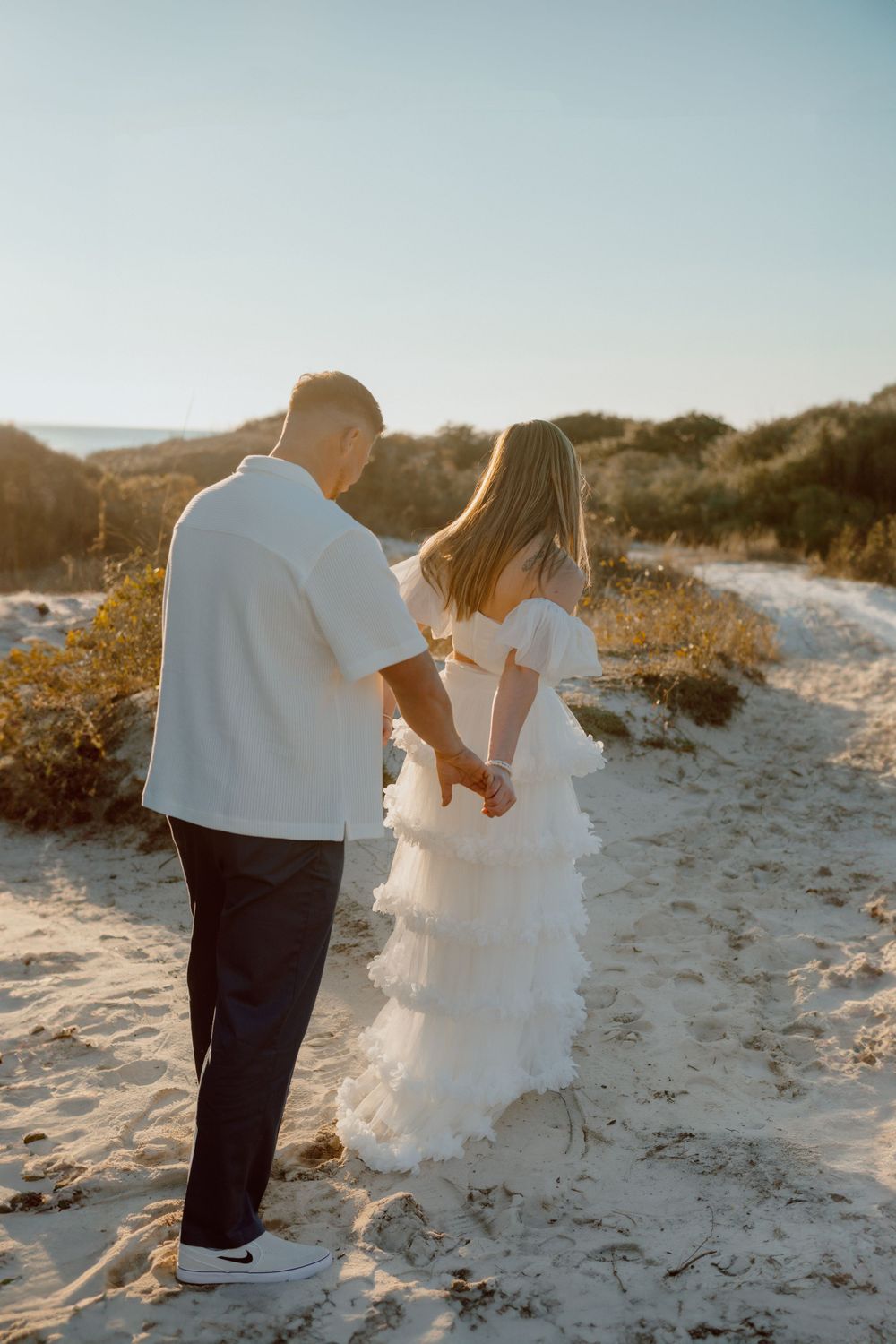 Couple holding hands on sandy beach path during golden hour.