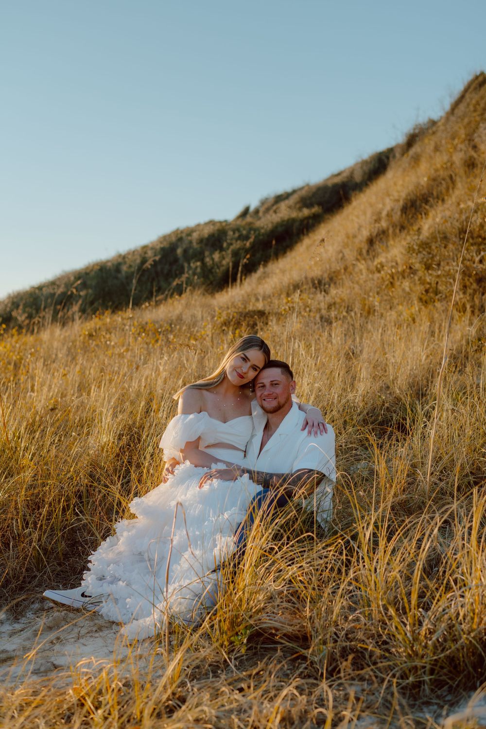Couple sitting together on sandy hill during sunset photoshoot.